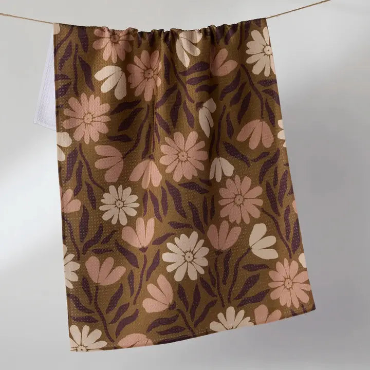 A brown kitchen towel with a retro floral pattern of pink, beige, and dark purple flowers, hanging from a string against a neutral gray background.