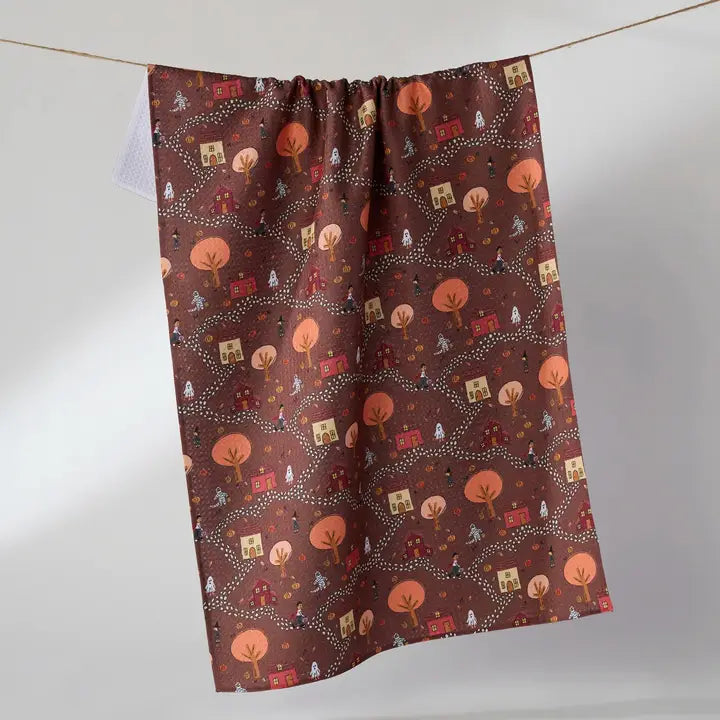 A rectangular kitchen towel with a brown background and an autumn-themed pattern of orange trees, red houses, and small white ghosts, hanging from a string against a light gray wall.