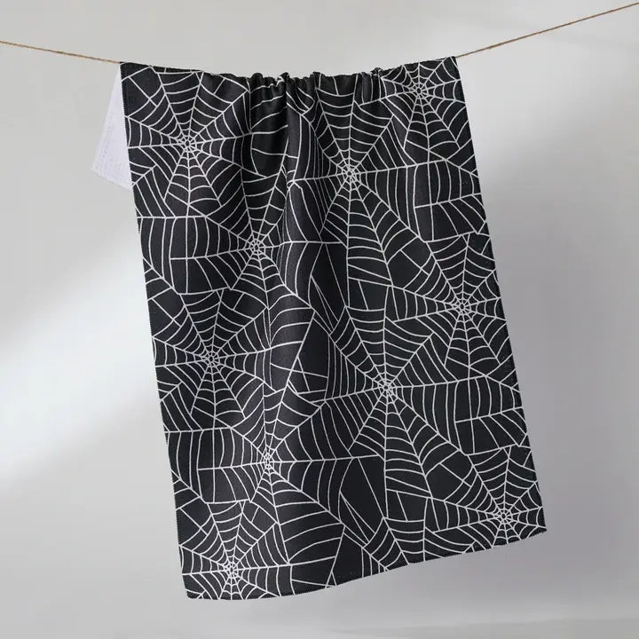 A black Geometry tea towel with a white spiderweb pattern, hanging from a thin wire against a light gray background.