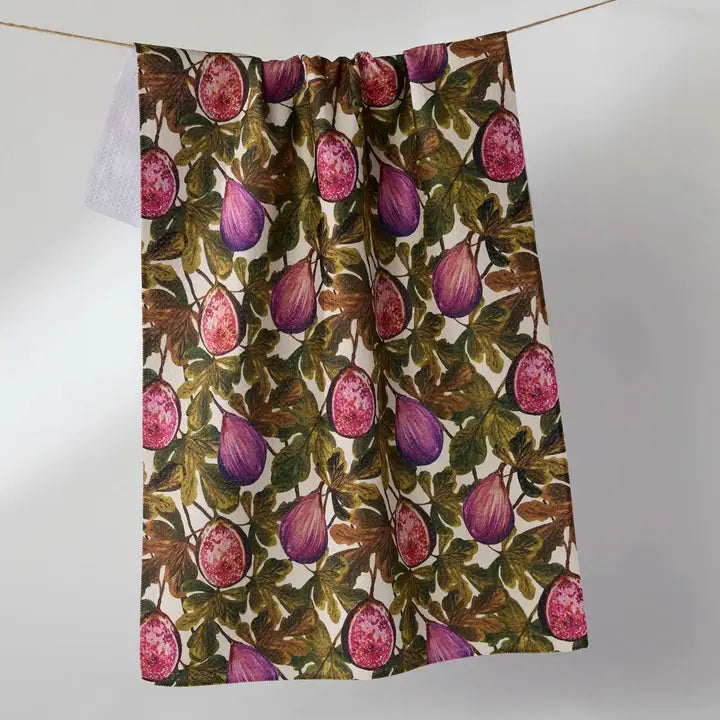 A kitchen tea towel with a vintage botanical print of purple figs and green leaves on a cream background, hanging from a string against a neutral gray background.