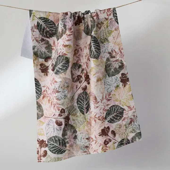 A fabric tea towel with a botanical leaf pattern in muted green, brown, and dusty pink, hanging from a thin cord against a light gray background.